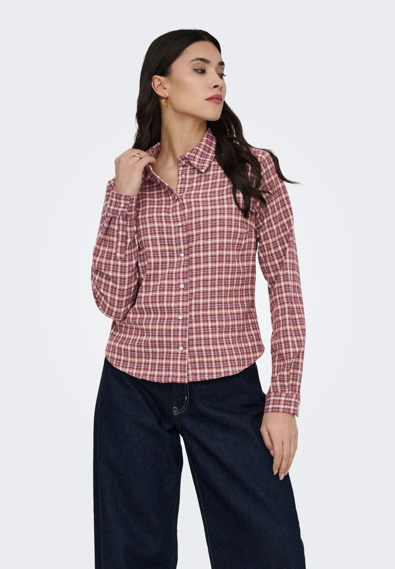 Femme portant une chemise à carreaux rouge et blanche boutonnée et un jean large bleu foncé, touchant son col et regardant sur le côté.