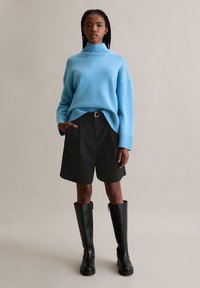 Lichtblauwe coltrui, losse pasvorm, geribde manchetten, gecombineerd met zwarte op maat gemaakte shorts en hoge zwarte laarzen. Neutrale achtergrond.