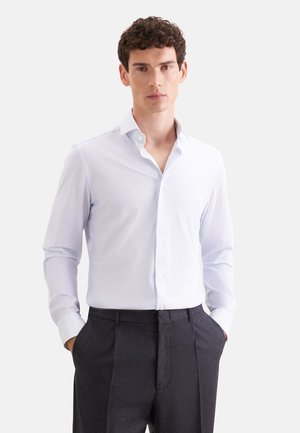 Jeune homme aux cheveux bouclés portant une chemise claire à motifs boutonnée et un pantalon sombre sur mesure, les mains dans les poches.