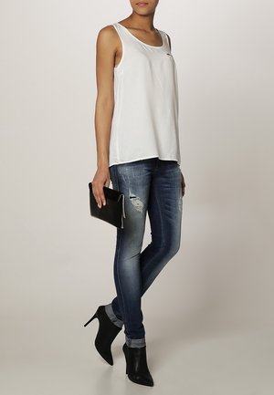 Débardeur blanc sans manches, jeans skinny bleu avec des détails déchirés, bottines noires à talons hauts et une pochette noire. Design simple et moderne.