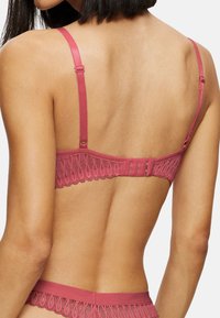 Rosa blonde bralette med justerbare stropper, blomstermønster langs båndet, og en krok-og-løkke lukking bak. Myk materiale tekstur.