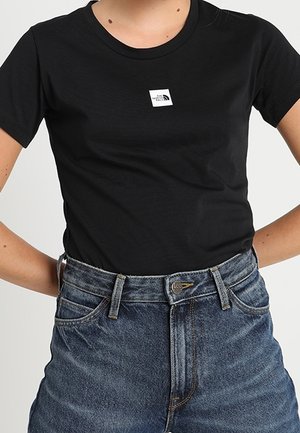 Person iført en tætsiddende sort The North Face t-shirt stoppet ned i højttaljede blå denimjeans med hænderne på hofterne.