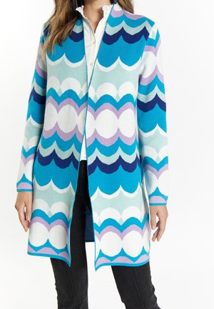Woman wearing a long, open-front cardigan with blue, white, purple, and teal scalloped wave patterns over a white shirt and black pants.