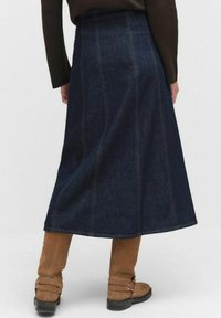 Denimowa spódnica maxi w ciemnoniebieskim kolorze, z pionowymi szwami, sylwetką w kształcie litery A i surowym wykończeniem; zestawiona z beżowymi botkami za kostkę z klamrami.