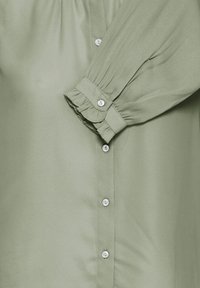 Light green blouse with gathered cuffs, white buttons down the front, and fabric ruffle detail at the wrist.