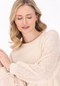 Pullover beige chiaro con maniche a palloncino in pizzo, dotato di una texture morbida, scollatura rotonda e polsini a costine.