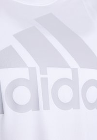 Camiseta deportiva blanca con un gran logotipo de adidas en gris claro en el frente. La tela parece suave con un ligero brillo.