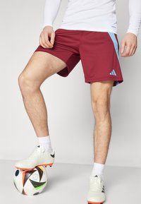 Bordeauxkleurige sportshorts met een lichtblauwe streep aan de zijkant, gemaakt van glad materiaal, gecombineerd met witte voetbalschoenen op een patroonbal.