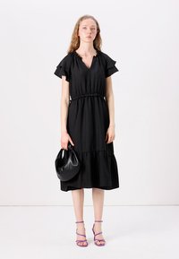 Black knee-length dress with short ruffled sleeves, an elastic waist, and tiered skirt. Paired with a black handbag and purple strappy heels.