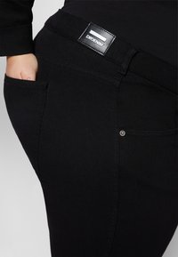 Jeans de ganga pretos com uma textura suave, apresentando um bolso traseiro e um patch de logo em pele na cintura. Detalhe de hardware minimalista em prata.
