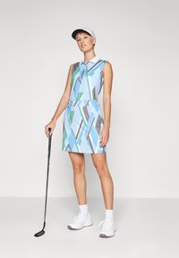 Golfové šaty bez rukávov, modrý základ, s geometrickými vzormi v zelenej a šedej farbe, doplnené šedou šiltovkou a kombinované s bielymi teniskami.