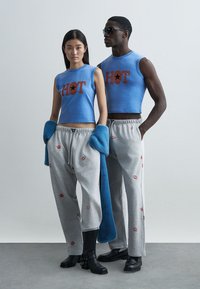 Top curto azul com a palavra "HOT" impressa em castanho, combinado com calças de fato de treino cinzentas com padrões de lábios vermelhos. A modelo usa botas pretas e um cachecol azul.