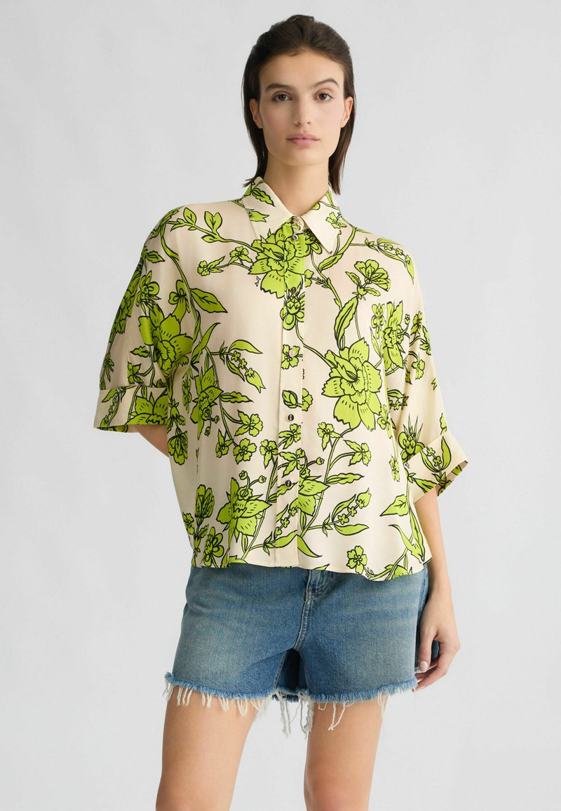 Kurzärmliges, beiges Blumenshirt mit lebendigem grünem Pflanzenmuster, mit durchgehender Knopfleiste und lockerer Passform. Kombiniert mit Jeansshorts.