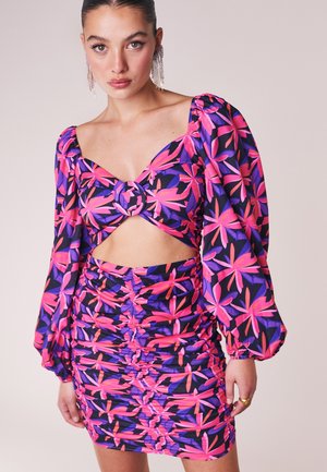Woman wearing a purple and pink floral long-sleeve crop top with ring detail and matching ruched mini skirt, standing against a plain background.
