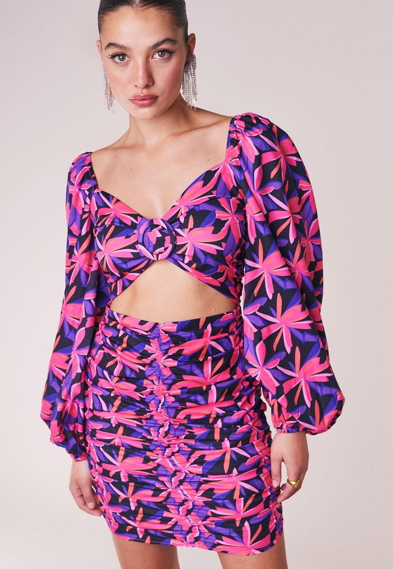 Femme portant un haut court à manches longues à fleurs violet et rose avec détail anneau et mini-jupe froncée assortie, debout devant un fond uni.