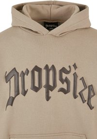 Beige Kapuzenpullover mit großem schwarzen gummierten Text "Dropsize" auf der Vorderseite, mit Kängurutasche und weicher Textur.