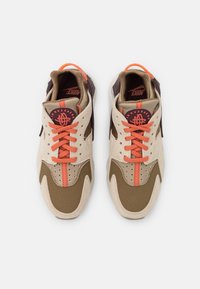 Ett par Nike Air Huarache sneakers i beige, brunt och orange med snörning framtill, synliga perforeringar och en märkesetikett på tungan.