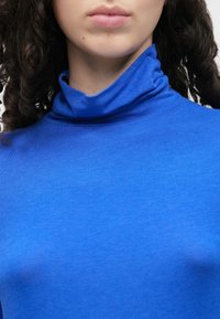 Zils augstas apkakles topiņš no mīksta auduma, kas cieši pieguļ apkaklei ar gludu tekstūru un minimālām šuvēm.