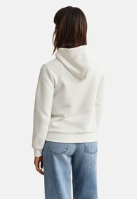 Femme portant un sweat à capuche blanc uni et un jean bleu clair, vue de dos sur un fond blanc uni.