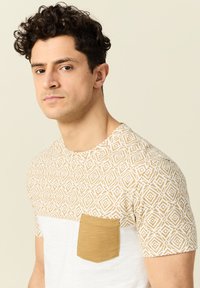 Lyhythihainen t-paita, jossa on geometrinen beige ja valkoinen kuvio yläosassa sekä kiinteä vaaleanruskea tasku rinnassa.