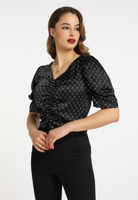 Schwarze Satinbluse mit V-Ausschnitt, geraffter Vorderseite und puffigen kurzen Ärmeln, verziert mit einem geometrischen Silbermuster. Kombiniert mit schwarzen Hosen.