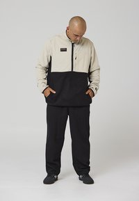 Giacca a pullover beige e nera con cerniera frontale, due tasche anteriori e una toppa con logo. Pantaloni neri e scarpe nere completano il look.