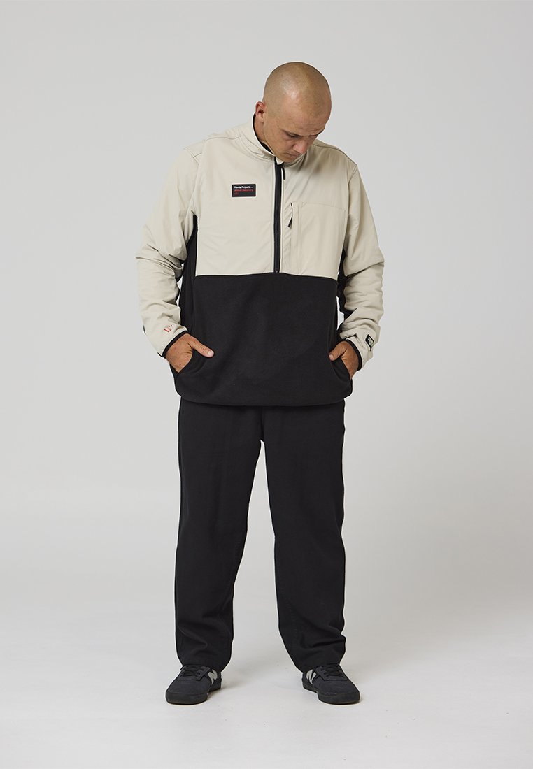 Giacca a pullover beige e nera con cerniera frontale, due tasche anteriori e una toppa con logo. Pantaloni neri e scarpe nere completano il look.