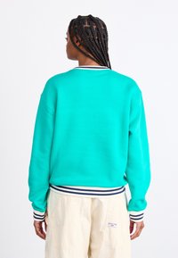 Τιρκουάζ φούτερ με ριμπ καβάλο και στρίφωμα σε μπλε και λευκές ρίγες. Το υλικό φαίνεται μαλακό, με σχέδιο χαλαρού κοψίματος.