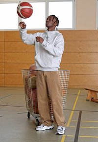 Una persona con una felpa grigia e pantaloni in velluto a coste beige bilancia un pallone da basket sulle punte delle dita in una palestra con pareti di legno e un carrello di metallo.