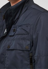 Mörkblå vadderad jacka med hög krage, flera framfickor, svarta tryckknappar och en ficka med dragkedja på sidan. Mjukt material.