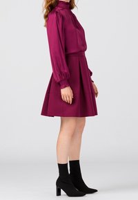 Mauve paita, jossa on kerätty kaulus ja pitkät hihat, yhdistettynä liehuvaan mauve-hameeseen. Yhdistetty mustiin sukka-saappaisiin ja sileään tekstiiliin.