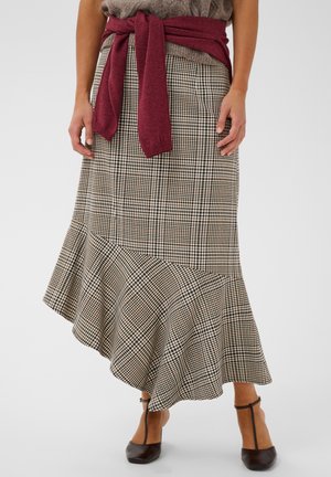 Femme portant une jupe asymétrique à carreaux taille haute avec un ourlet à volants, un pull bordeaux noué autour de la taille, et des talons noirs à bride en T.