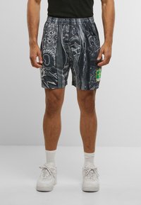 Mand med sort og grå mønstrede shorts med en gul og grøn lap, hvide sokker og hvide sneakers, der står imod en ensfarvet baggrund.