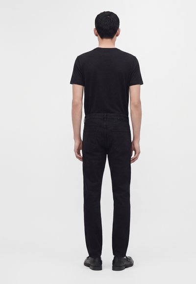 Homme debout de dos, vêtu d'un t-shirt noir, d'un jean noir et de chaussures noires sur un fond clair uni.