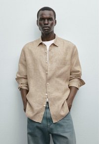 Light brown linen shirt with a button-down front, single chest pocket, and rolled sleeves, paired with blue denim jeans.
