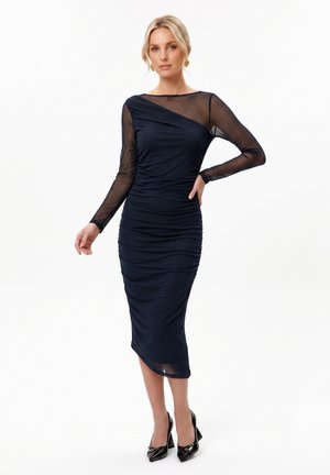 Femme portant une robe midi bleu marine ajustée avec des manches longues transparentes et des chaussures à talons noires, debout sur fond blanc.