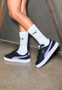 Baskets en daim noir avec une bande violette, une semelle en caoutchouc blanche et des détails de logo dorés, associées à des chaussettes blanches côtelées avec un logo noir.
