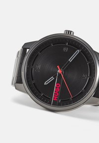 Reloj de metal negro con esfera texturizada, marcadores de hora plateados y detalles en rojo. Cuenta con una pulsera de acero inoxidable negro y una llamativa marca "HUGO".