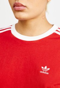 Rød bomull T-skjorte med hvit ribbestrikket krage, tre hvite striper på skuldrene og liten hvit Adidas-logo på brystet.