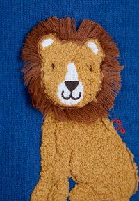 Gestickter Plüsch-Löwe mit goldenem Körper und braunem Mähnen auf einem blauen gestrickten Hintergrund. Verfügt über strukturierte Färbung und gestickte Gesichtszüge.