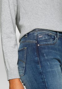 Grå ribbad tröja kombinerad med mörkblå denimjeans med en subtil tvättad textur och en liten logotyp nära bakfickan.