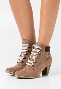 Stivaletti in suede marrone con punta a punta, dotati di lacci a strisce bianche e marroni, occhielli in metallo e tacco impilato.