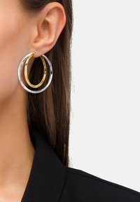 Närbild på ett öra med en dubbel hoop-örhänge, en i guld och en i silver, mot brun hår och en svart krage.