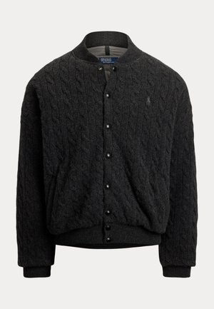 Jeune homme portant une veste noire en maille torsadée, une écharpe à carreaux gris, un pantalon gris foncé, des baskets noires et des lunettes de soleil rondes, debout les mains dans les poches.