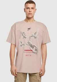 Lyserød oversized t-shirt lavet af bomuld, med et grafisk design af duer og kæder, med teksten "FREEDOM" trykt i rød.