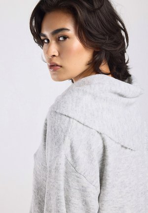 Persoon met halflang donker haar draait het hoofd, draagt een lichtgrijze textuur hoodie tegen een effen lichte achtergrond.