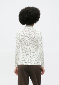 Persona con cabello rizado, vistiendo una blusa larga de encaje floral blanca y pantalones marrón oscuro, mostrada de espaldas contra un fondo liso.