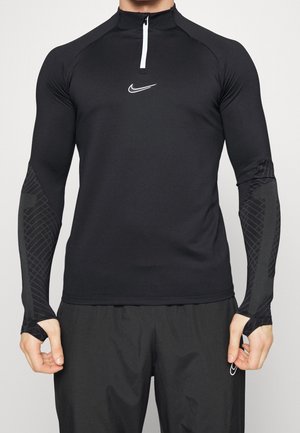 Homme portant un haut de sport noir ajusté à manches longues avec une fermeture éclair blanche et un petit logo blanc sur la poitrine, associé à un pantalon noir.