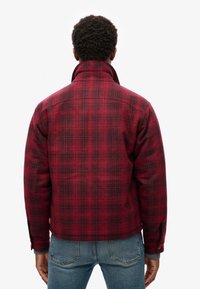 Chaqueta a cuadros en rojo y negro con cuello alto y puños abotonados; el material parece texturizado, con un corte relajado y una línea de dobladillo corta.