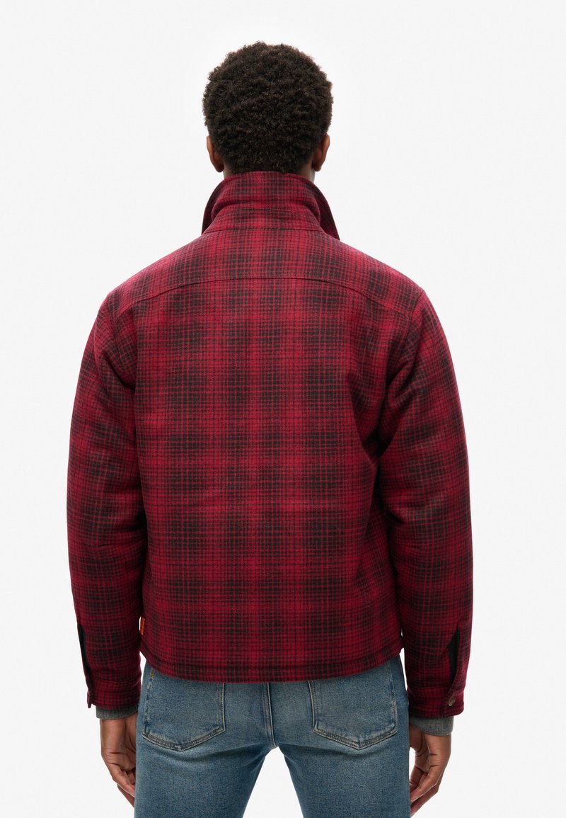 Rot-schwarz karierte Jacke mit hohem Kragen und geknöpften Manschetten; das Material wirkt strukturiert, mit einer lockeren Passform und einer verkürzten Saumlinie.